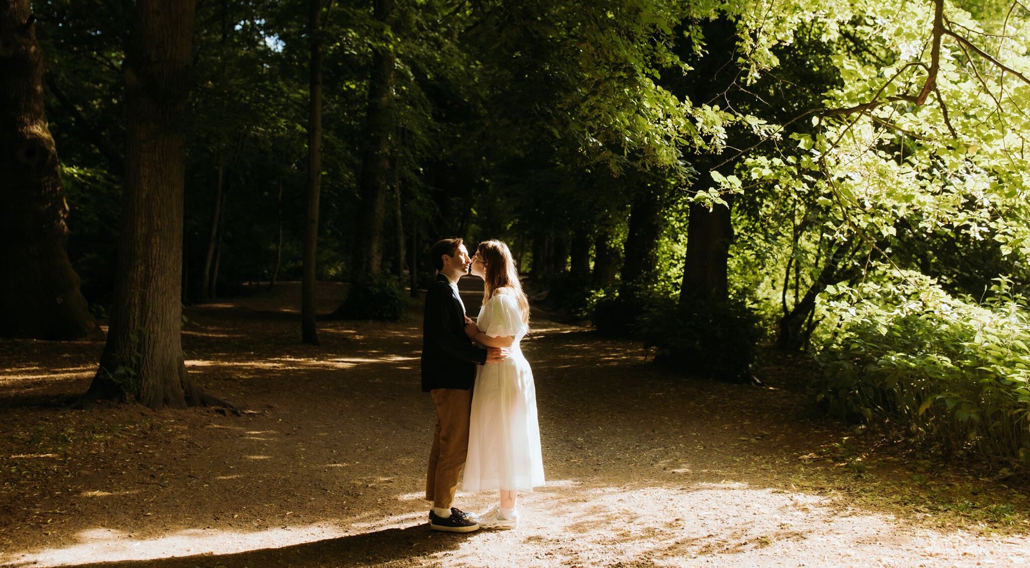 A couple kiss in a bright patch of light on a wooded path, amongst the trees of Hampstead Heath